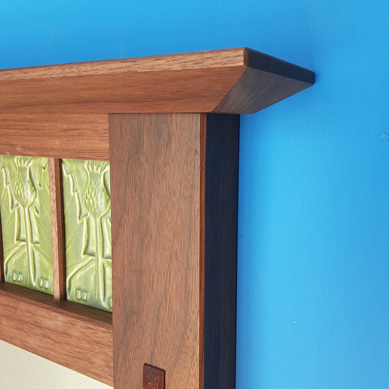 Puede incluir: Una repisa de chimenea de madera con un acabado de veta de madera marr&oacute;n oscuro y un estante superior horizontal. La estructura incorpora paneles rectangulares insertados con azulejos de cer&aacute;mica verde claro con un motivo de cardo.