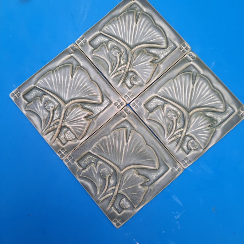 May include: A set of three square ceramic tiles arranged in a diamond formation. Each tile features a raised, embossed ginkgo leaf design in a muted, metallic bronze-grey finish with small square grid patterns at the corners of each tile.