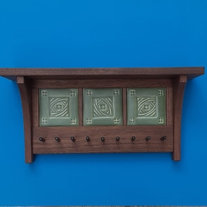 May include: A wooden wall shelf with three green ceramic tiles featuring a geometric design. The shelf has ten hooks for hanging items.