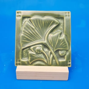 May include: A square, sage green ceramic tile featuring a raised relief design of ginkgo leaves. The tile has a decorative border with small square accents in each corner and is displayed resting on a rectangular, light-colored wooden stand.