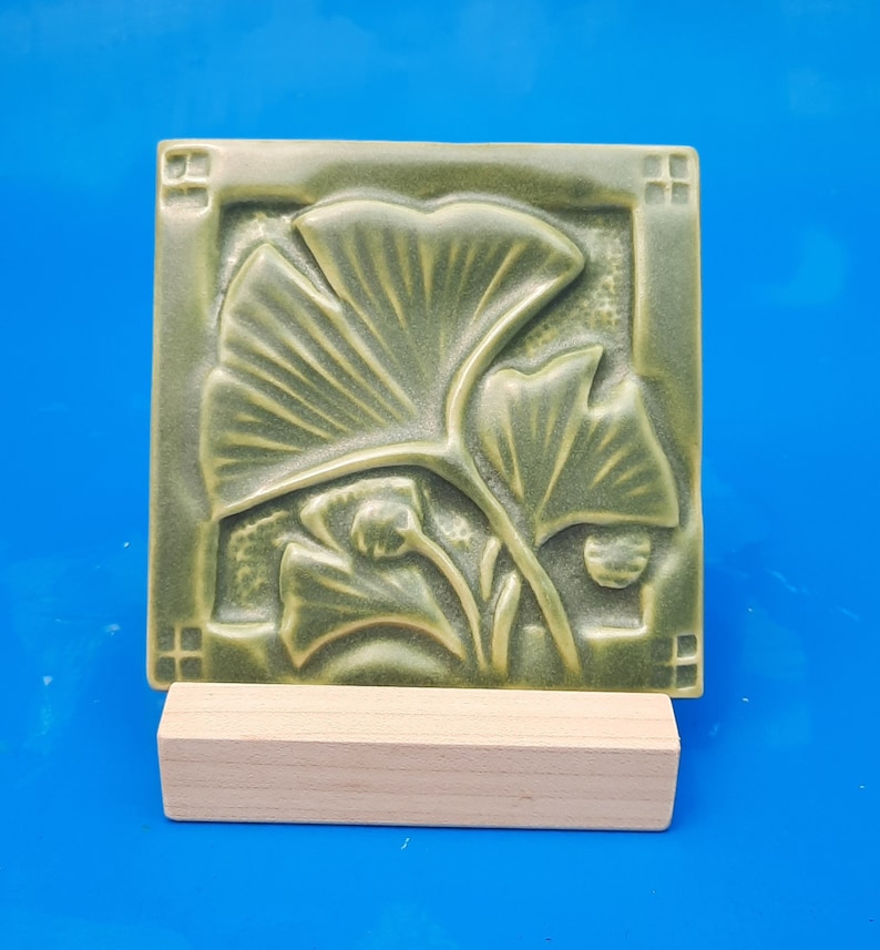 May include: A square, sage green ceramic tile featuring a raised relief design of ginkgo leaves. The tile has a decorative border with small square accents in each corner and is displayed resting on a rectangular, light-colored wooden stand.