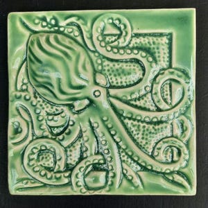 Op de afbeelding: Vierkante keramische tegel met een reliëfontwerp van een octopus in groentinten. De octopus heeft een grote kop en meerdere tentakels met zuignappen. De tegel heeft een glanzende afwerking en een decoratieve rand.