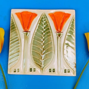 May include: A square ceramic tile with a floral design featuring two orange flowers and a central green leaf. Two yellow California poppies flank the tile on a blue background. The tile is likely handmade.