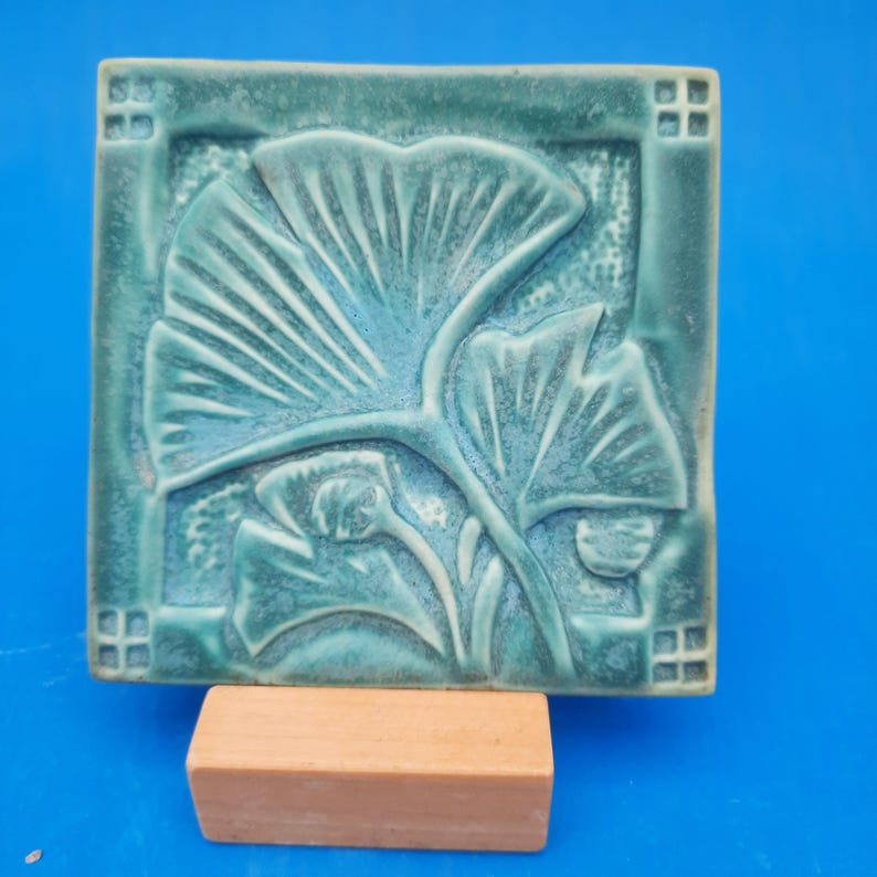 May include: A square ceramic tile featuring a raised relief design of ginkgo leaves in shades of teal and green. The tile has a textured border with small square grid patterns in each corner and rests on a rectangular wooden block stand.