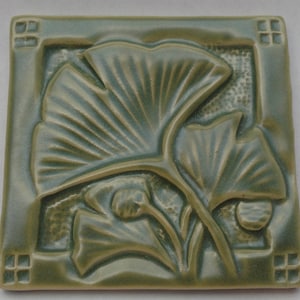 May include: A square ceramic tile in a muted sage green glaze featuring a raised relief design of ginkgo leaves. The border includes small square cutouts at each corner, and the background texture behind the leaves is subtly stippled.