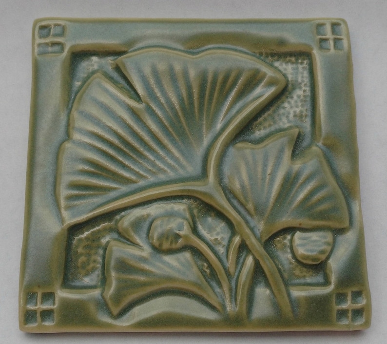 May include: A square ceramic tile in a muted sage green glaze featuring a raised relief design of ginkgo leaves. The border includes small square cutouts at each corner, and the background texture behind the leaves is subtly stippled.