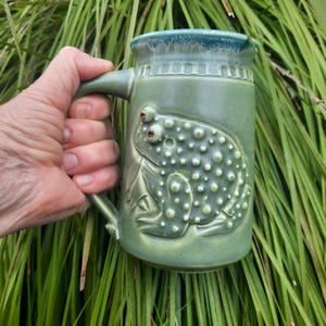 May include: A green ceramic mug featuring a raised frog design. The frog has brown eyes and a textured body.