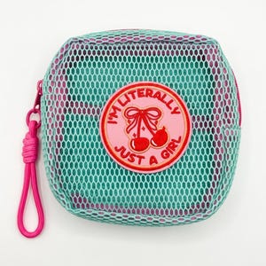 May include: A square zippered pouch with a teal mesh outer layer over a purple fabric base. A circular pink patch is centered on the front, featuring an embroidered red cherry graphic with a bow and the text I'M LITERALLY JUST A GIRL.