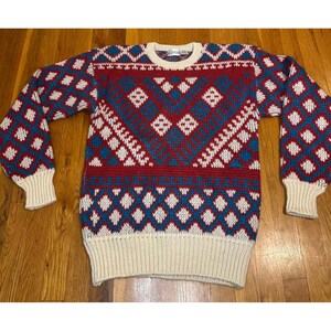 Könnte beinhalten: Ein langärmeliger Strickpullover mit einem geometrischen Rautenmuster in Rot, Blau und Weiß. Das Kleidungsstück hat einen Rundhalsausschnitt, gerippte Bündchen und einen gerippten Saum in einfarbigem Creme. Das Muster bedeckt Brust und Ärmel.