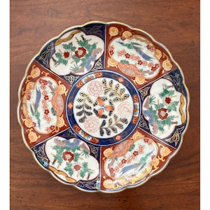 May include: A round ceramic plate with a scalloped edge featuring a central circular medallion with floral motifs. The plate is divided into six alternating panels of white and rust-red, decorated with painted birds, flowers, and gold accents.