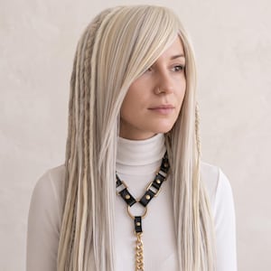 May include: A long, straight, platinum blonde wig featuring side-swept bangs and multiple thin, textured braids integrated into the hair. The wig is worn over a white ribbed turtleneck sweater and a black leather harness with gold-toned metal hardware.