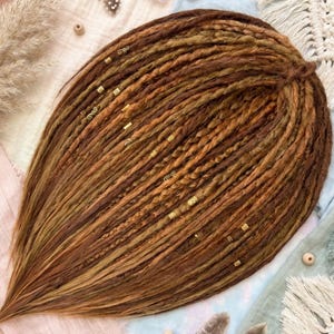 Op de afbeelding: Een set bruine en goudbruine dreadlocks, versierd met kleine goudkleurige kralen. De dreadlocks zijn in een waaier gerangschikt, met een mix van lichte en donkere tinten. De afbeelding is van bovenaf genomen.