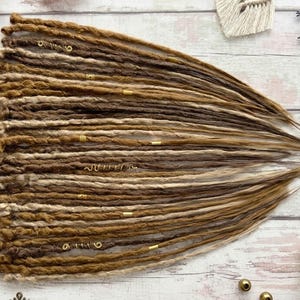 May include: A bundle of handmade dreadlocks in varying shades of brown and blonde. The dreadlocks are styled with small gold-colored metal accents. The hair extensions are arranged on a light-colored wooden surface.