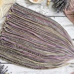 May include: A bundle of braided hair extensions in shades of lavender, gray, and beige. The extensions are arranged in a fan shape, with the braids neatly aligned. The hair is styled with a gradient effect, transitioning from darker to lighter tones.