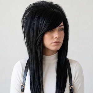 Puede incluir: Una peluca negra larga y lisa con gran volumen en la coronilla y flequillo lateral. El cabello está escalonado, llega más allá de los hombros y se lleva sobre una prenda blanca de cuello alto con tiras negras que tienen hebillas doradas.