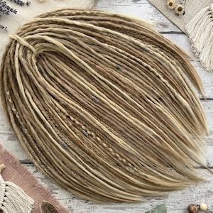 May include: A bundle of blonde and light brown synthetic dreadlocks, arranged in a fan shape. The dreadlocks are a mix of solid and braided strands, with small silver-colored beads interspersed throughout. The hair is styled on a neutral-toned surface.