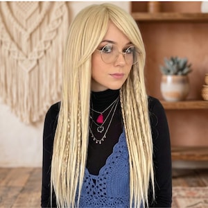 May include: A woman with long, straight blonde hair and side-swept bangs, wearing round eyeglasses, a black turtleneck, and a blue crochet top. She is also wearing multiple silver necklaces, one with a pink tassel.