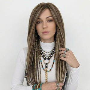 May include: A woman wearing a white turtleneck shirt, adorned with multiple necklaces featuring turquoise and wooden beads, and a black leather harness. Her fingers are adorned with rings, and she has long, light brown hair with dreadlock accents.