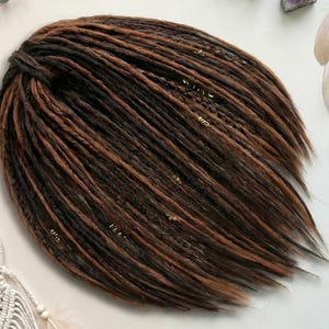 Op de afbeelding: Een bundel bruine en zwarte dreadlocks, met een mix van donker- en lichtbruine strengen. De dreadlocks zijn versierd met kleine, goudkleurige kralen. Het haar is in een nette, georganiseerde opstelling gestyled.