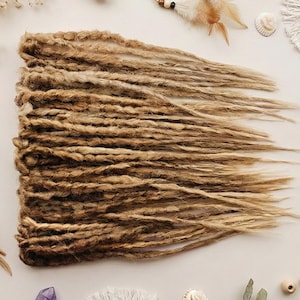 Peut inclure: Un ensemble de dreadlocks bruns clairs et blonds, disposés sur une surface neutre. Les dreadlocks ont un aspect naturel et texturé. L'image est agrémentée d'éléments décoratifs tels que des cristaux, des plumes et des coquillages, suggérant une esthétique bohème ou alternative.