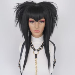 Puede incluir: Un arnés de cuero negro con herrajes dorados y una cadena dorada. El arnés se lleva sobre un cuello alto blanco. El maniquí tiene cabello negro con flequillo y lleva maquillaje.