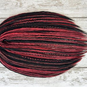 May include: A bundle of red and black dreadlocks, arranged in an oval shape. The dreadlocks are a mix of solid red and black strands, with some sections appearing a darker shade of brown. The background is a light-colored, weathered wooden surface.