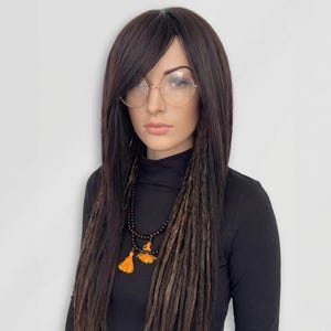 May include: A person wearing a black turtleneck shirt, round gold-rimmed glasses, and a long dark brown wig with side bangs. The wig has long, dark brown dreadlocks with lighter brown highlights. A black beaded necklace with orange tassels is also visible.