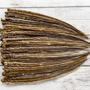 May include: A collection of brown and blonde dreadlocks, some with gold-colored metal accents. The dreadlocks are arranged on a light-colored wooden surface, showcasing a natural, textured appearance. The color palette includes various shades of brown and blonde.