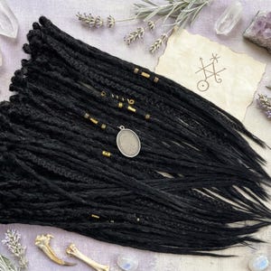 May include: A bundle of black dreadlocks with gold-colored beads and a silver pendant. The dreadlocks are arranged on a light purple surface, with sprigs of lavender, crystals, and a parchment with a symbol.