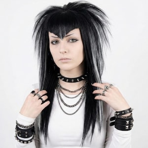 Puede incluir: Una persona con una camisa blanca de manga larga, joyas negras y plateadas y pulseras de cuero negro. Las joyas incluyen un collar con tachuelas, collares de cadena en capas, anillos y pulseras. El cabello es negro con mechones blancos.