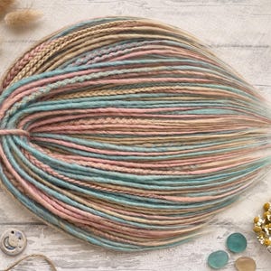 May include: A bundle of pastel-colored synthetic dreadlocks in shades of pink, blue, and beige. The dreadlocks are arranged in a fan shape, with a braided section at the top. The color palette is soft and feminine, suitable for a bohemian or alternative style.