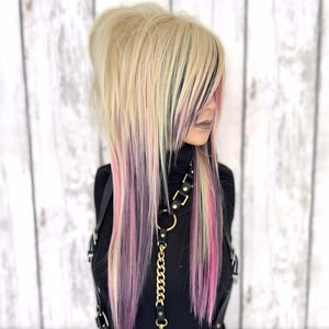 May include: A woman with long, straight blonde hair with pink and black streaks. She is wearing a black mock neck shirt and a black leather harness with a gold chain.