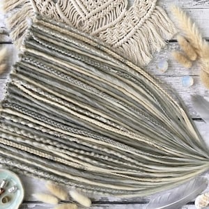 May include: A set of synthetic hair extensions featuring a blend of light gray, ash, and cream-colored dreadlocks and braids. The strands are adorned with small silver-toned metal beads and cuffs, arranged in a fan-like pattern on a flat surface.