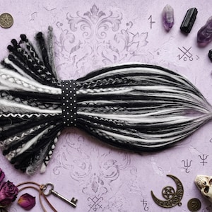 May include: A black, white, and gray dreadlock hair extension piece, secured with a black polka dot band. The arrangement is on a lavender surface with decorative elements including crystals, coins, a key, and a skull.