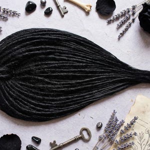 May include: A large bundle of black dreadlocks, with a textured appearance, is the central focus. The dreadlocks are surrounded by decorative items such as dried lavender sprigs, black rose petals, antique keys, and small black stones.