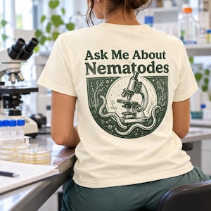 Könnte beinhalten: Ein cremefarbenes Kurzarm-T-Shirt mit einer dunkelgrünen Grafik auf der Rückseite. Das Design zeigt ein Mikroskop, wurmartige Nematoden und den Text "Ask Me About Nematodes", der in einer fetten, dunkelgrünen Schrift über dem Bild steht.