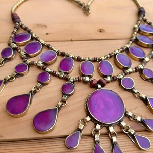 May include: A multi-layered statement necklace featuring numerous oval and teardrop-shaped purple cabochons set in brass metal frames. The design includes small brown wooden beads and brass spacers strung along the chain, with dangling purple pendants.