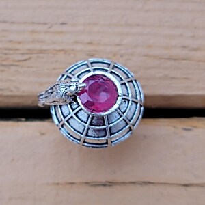May include: A silver ring featuring a round, ruby-colored gemstone set within a cage-like design. A detailed bird head is sculpted on the side of the ring. The ring is set against a wooden background.