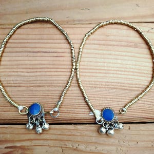 A pair of gold-colored metallic bead anklets with blue stone charms and dangling silver bells resting on a wooden surface.