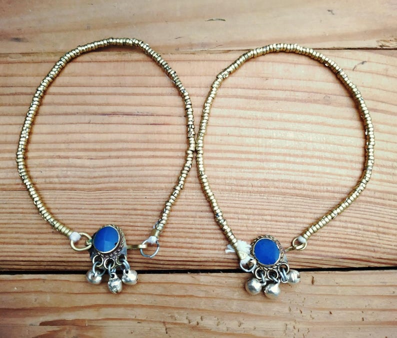 A pair of gold-colored metallic bead anklets with blue stone charms and dangling silver bells resting on a wooden surface.