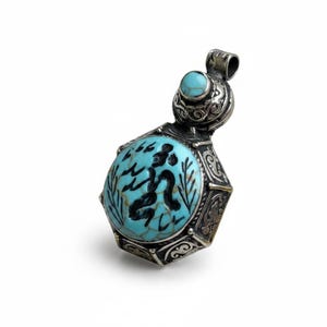May include: A silver pendant featuring a large, round, light blue turquoise stone with black matrix and a dark etched figure. The metal setting is hexagonal with intricate scrollwork, topped by a smaller round turquoise stone and a bail.