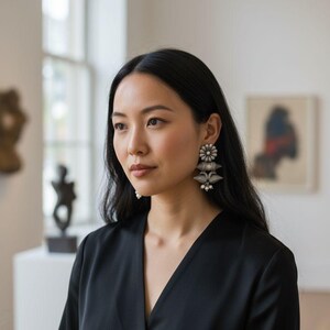 Op de afbeelding: Een vrouw draagt zilveren oorbellen met een bloemmotief en bungelende elementen. De oorbellen zijn het middelpunt, tegen een zwarte top. De achtergrond is een galerij met kunstwerken en natuurlijk licht.