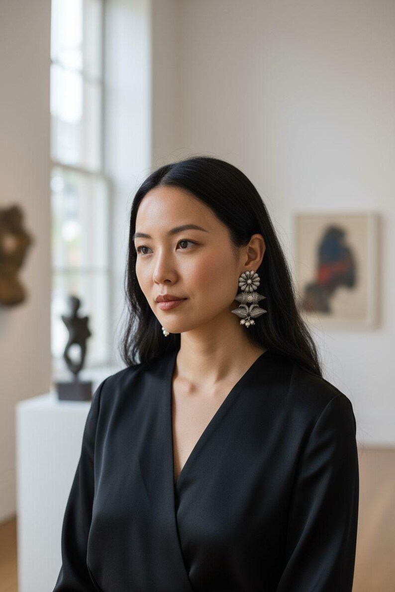 Op de afbeelding: Een vrouw draagt zilveren oorbellen met een bloemmotief en bungelende elementen. De oorbellen zijn het middelpunt, tegen een zwarte top. De achtergrond is een galerij met kunstwerken en natuurlijk licht.