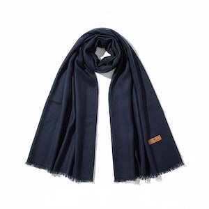 May include: A navy blue scarf with a fringed edge and a small, rectangular brown leather tag. The scarf is draped on a white background. The scarf is made of a soft material.