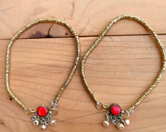 May include: A pair of gold-colored anklets with a beaded chain design. Each anklet features a central red stone and dangling silver-colored bells. The anklets are displayed on a wooden surface.
