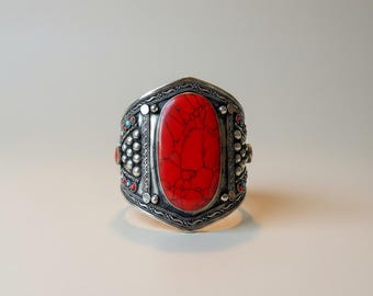May include: A silver cuff bracelet with a large, oval, red stone as the centerpiece. The bracelet has intricate detailing and is adorned with smaller turquoise and red stones. A bold statement piece.
