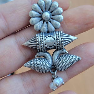 Op de afbeelding: Zilverkleurige oorbellen met een bloemvormige bovenkant, een geometrisch middenstuk en een pauwvormige onderkant. De oorbellen hebben een bungelende cluster van kleine, witte parels. Het ontwerp is ingewikkeld en gedetailleerd, met een vintage uitstraling.