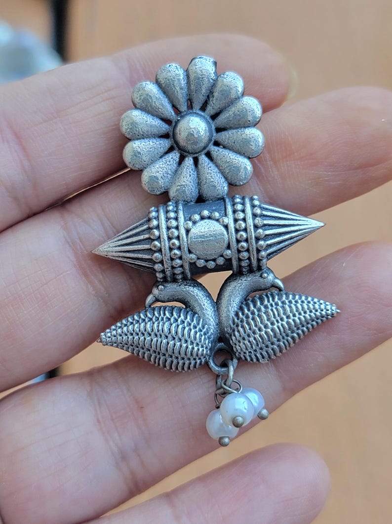Op de afbeelding: Zilverkleurige oorbellen met een bloemvormige bovenkant, een geometrisch middenstuk en een pauwvormige onderkant. De oorbellen hebben een bungelende cluster van kleine, witte parels. Het ontwerp is ingewikkeld en gedetailleerd, met een vintage uitstraling.