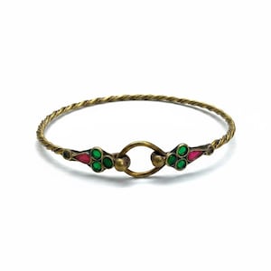 Puede incluir: Una pulsera rígida de metal dorado con una banda de alambre trenzado. El punto focal central consiste en un anillo circular flanqueado por dos elementos decorativos, cada uno incrustado con tres piedras verdes pequeñas y una piedra roja en forma de gota.