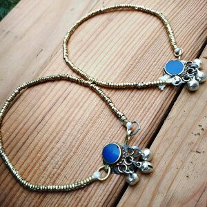 Two gold-tone beaded anklets with blue cabochons and hanging bell charms displayed diagonally on wooden planks.